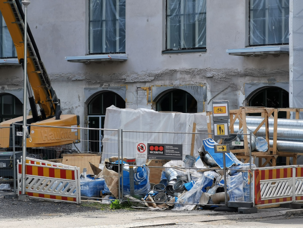 Baustelle mit einem großen Gebäude im Hintergrund, einem Kran, einem Müllhaufen, Absperrungen, Schildern, Rohren und verstreuten Gegenständen im Vordergrund.