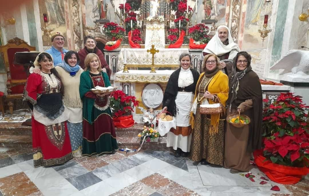 Eine Gruppe von Menschen versammelt sich vor einer Kirche mit Körben und Tellern, im Hintergrund sind Krippen-Schmuck wie Blumenbouquets, Statuen, Kerzen und Gemälde zu sehen.