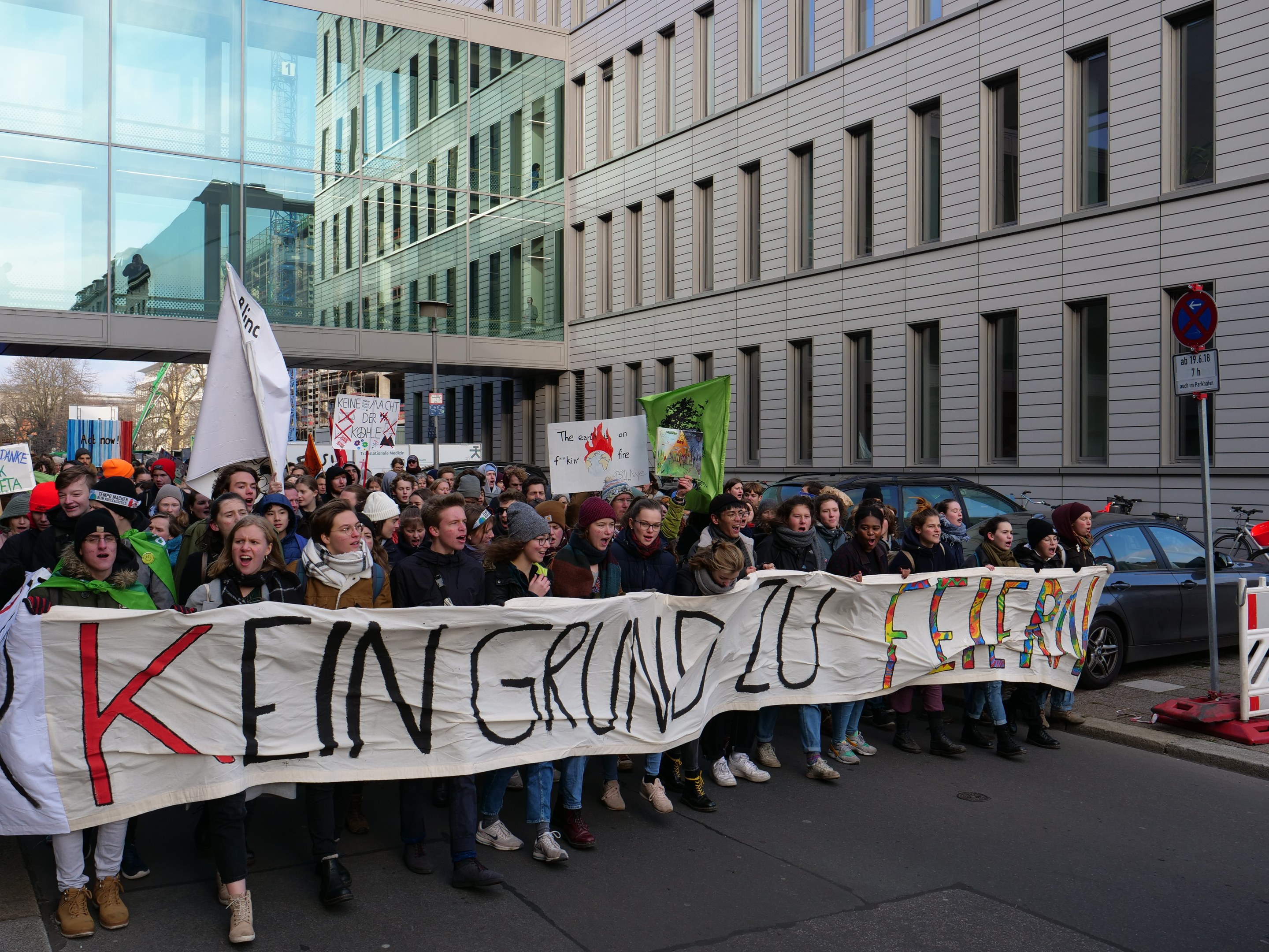 Eine Gruppe von Menschen mit einem Transparent mit der Aufschrift "Kein Sparen auf Kosten der Armen" demonstriert auf einer Straße mit Gebäuden, Bäumen und Fahrzeugen im Hintergrund unter einem klaren blauen Himmel.