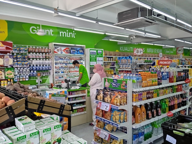 Ein Ladeneinrichtungsbild mit Regalen voller verschiedener Lebensmittel, darunter Flaschen, Schachteln und Früchte, sowie an der Decke montierte Tafeln, Lampen und ein Projektor, was auf einen neu eröffneten Mini-Supermarkt in Malaysia hinweist.