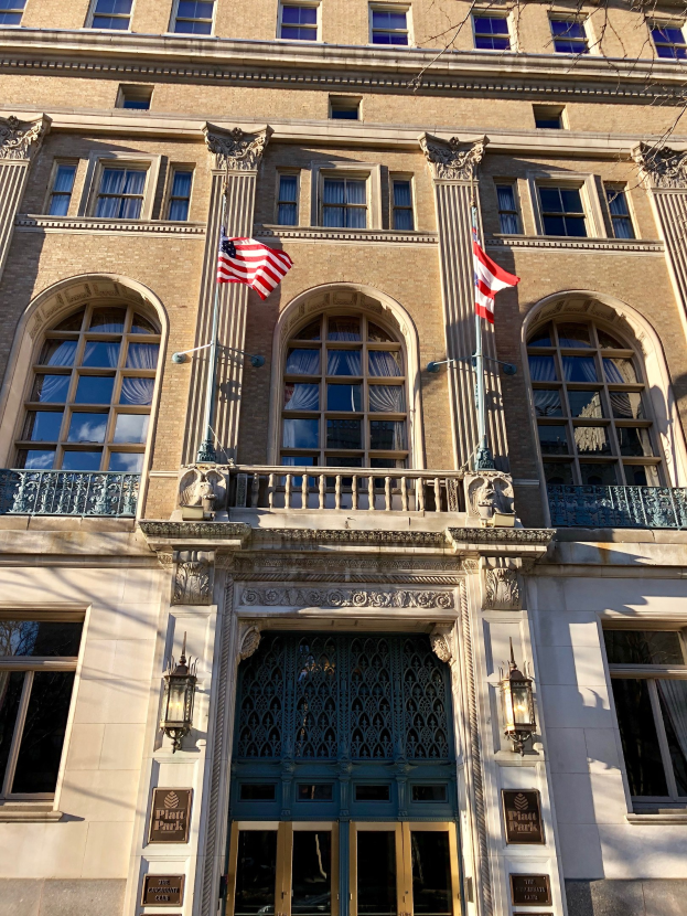 Großes Gebäude mit Glasfenstern, -türen, -säulen, -geländern, -leuchten, Texttafeln und Flaggen, identifiziert als Plaza Hotel in New York City vor einem Himmelhintergrund.