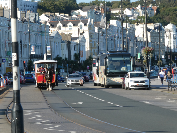 Eine belebte Stadtstraße mit zahlreichen Fahrzeugen, Fußgängern, hohen Gebäuden, Verkehrsampeln, Laternenmasten, Texttafeln und Bäumen unter einem klaren blauen Himmel.