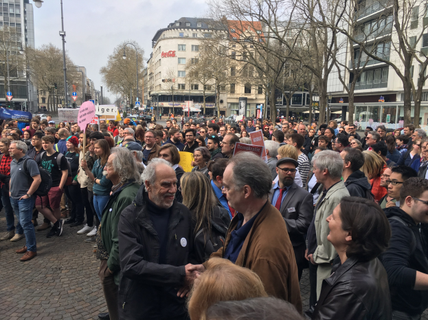 Eine große Gruppe von Menschen steht vor einer Menge mit Schildern, mit Fahrzeugen, Bäumen, Laternenmasten und Gebäuden im Hintergrund, wahrscheinlich während einer Demonstration in Berlin, Deutschland.