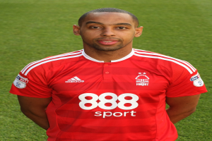 Ein Mann in einem roten T-Shirt steht selbstbewusst auf einem Fußballfeld mit saftig grünem Gras und zeigt einen entschlossenen Gesichtsausdruck.