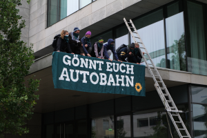 Eine Gruppe von Menschen steht auf dem Dach eines Gebäudes, hält eine Fahne, mit einem Baum links und Glasfenstern im Hintergrund, wahrscheinlich protestierend im Support der deutschen Regierung.