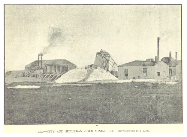 Schwarzes und weißes Foto einer Stadt und eines suburbanen Goldbergbauunternehmens mit Gebäuden, Polen und Rauch im Hintergrund, mit Text unten.