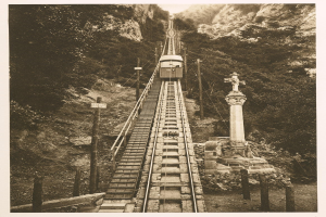 Ein altes Schwarz-Wei├č-Foto eines Zuges, der eine steile Steigung hinauff├Ąhrt, mit einem Pfahl auf der rechten Seite und B├Ąumen im Hintergrund und Text unten.