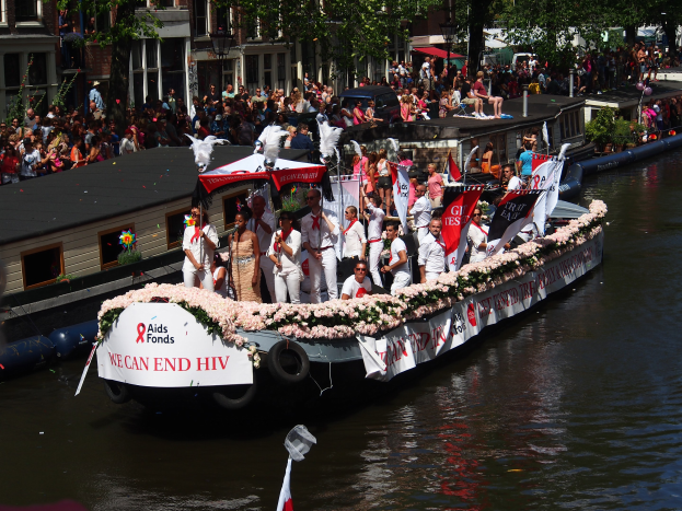 Eine Gruppe von Menschen auf einem Boot, das mit Flaggen und Blumen geschmütckt ist, bei der Amsterdam-Rot-Kreuz-Parade, mit Gebäuden, Bäumen und einem Laternenpfahl im Hintergrund.