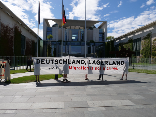 Eine Gruppe von Menschen mit einer Fahne mit der Aufschrift "Deutschland, Lagerland Migration ist kein Verbrechen" vor einem Gebäude mit Fenstern, umgeben von Gras, einem Metallzaun, Pflanzen, Bäumen und Fahnenstangen.