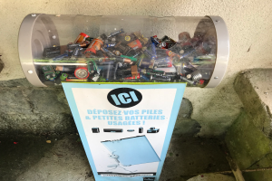 Ein Mülltonnen voller Batterien auf einem Gehweg, mit einem Plakat mit Text an der Wand dahinter und einer Treppe auf der rechten Seite.