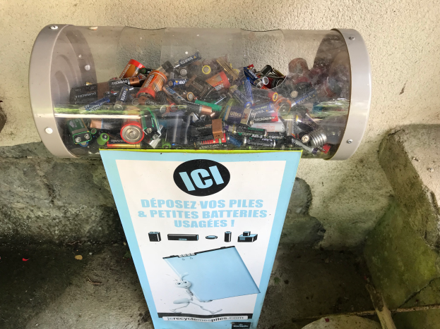 Ein Mülltonnen voller Batterien auf einem Gehweg, mit einem Plakat mit Text an der Wand dahinter und einer Treppe auf der rechten Seite.