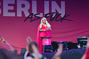 Eine Frau in einem pinken Kleid steht an einem Podium und hält ein Mikrofon und Papier in der Hand, mit einem 'Berlin as Fuck'-Plakat im Hintergrund, während sie eine Menge anspricht.