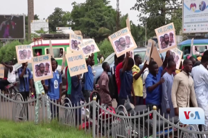 Gruppe von Menschen mit Schildern vor einem Zaun, die gegen ein Regierungsverbot von Video-Konferenzen protestieren, mit Fahrzeugen, Bäumen und einem Bildschirm im Hintergrund.