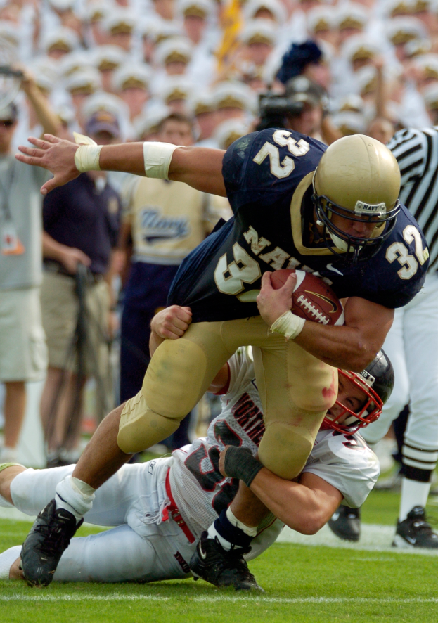 Ein Fussballspieler in einem Sportdress und Helm wird von einem anderen Spieler auf dem Feld getackelt, während andere Spieler und Zuschauer im Hintergrund zu sehen sind.