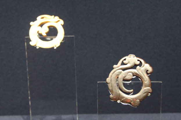 Goldene runde Ohrringe mit detaillierten Designs in einer durchsichtigen Glasvitrine präsentiert.