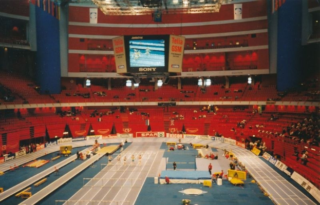 Großes Hallenstadion mit Zuschauern bei einer Leichtathletikveranstaltung, mit Sitzbereichen, Stehplätzen, Texttafeln, Beleuchtung und einem Bildschirm oben.