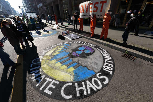 Eine Gruppe von Menschen demonstriert auf einer Straße, einige tragen Masken und halten Schilder mit der Aufschrift "Klimachaos", mit verstreuten Flaschen und Gegenständen auf dem Boden, vor einem Hintergrund von Gebäuden, Laternen und einem klaren blauen Himmel.