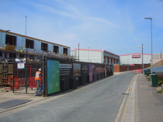 Eine Straße mit einem Gehweg, Laternen, Mülltonnen, Pflanzen und einem Zaun, im Vordergrund eine Baustelle und im Hintergrund Gebäude, einen Turm und den Himmel.