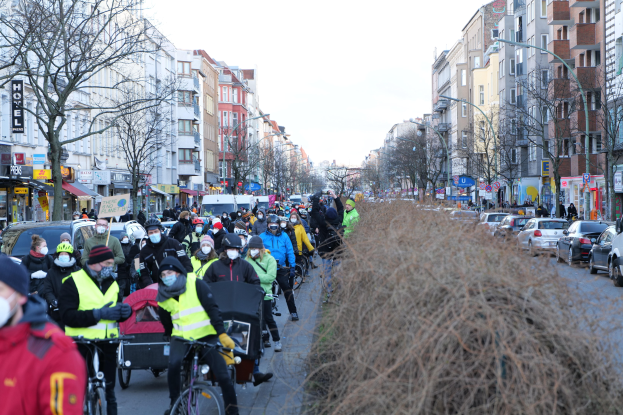 Eine große Gruppe von Menschen mit Masken und Sicherheitswesten fährt auf Fahrrädern eine von Bäumen gesäumte Straße mit Gebäuden, Laternenmasten und Texttafeln entlang, während Fahrzeuge die Straße teilen und trockenes Gras die rechte Seite säumt unter einem klaren blauen Himmel.