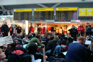 Eine große Gruppe von Menschen versammelt sich in einem Flughafen, einige sitzen mit Taschen und Papieren, andere stehen, mit Texttafeln, Schaufensterpuppen in Kleidern und Deckenlampen im Hintergrund, was auf eine Demonstration hindeutet.