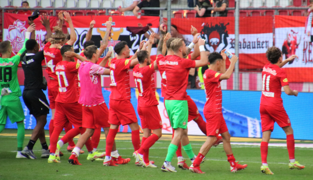 Gruppe von Fußballspielern, die ein Tor feiern, einige halten Flaschen und andere klatschen, mit einem Zaun, Bannern, Metallstangen, Stühlen und einer Decke mit Deckenleuchten im Hintergrund.