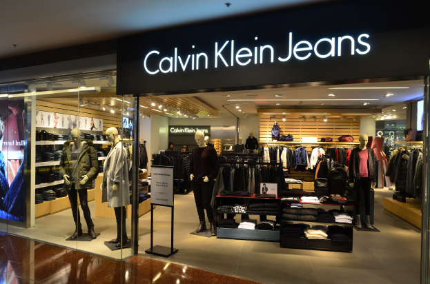 Innenansicht eines Calvin Klein Jeans-Geschäfts in einem Einkaufszentrum, das Schaufensterpuppen in aktuellen Kleidungsstilen, aufgehängte Kleidung, eine Werbetafel, einen digitalen Bildschirm und eine Deckenbeleuchtung zeigt.