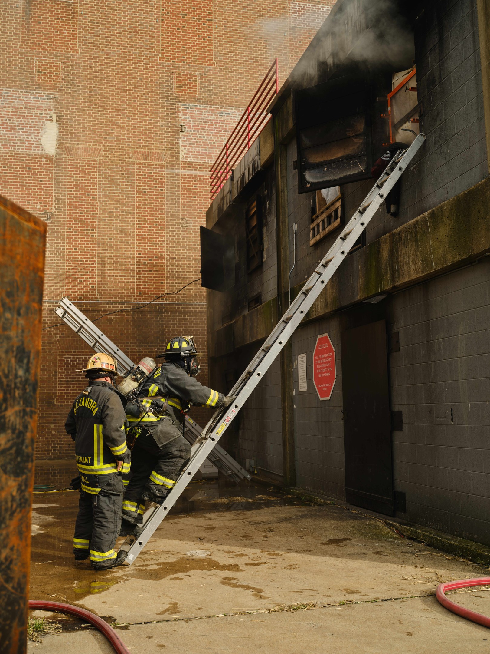 Feuerwehrleute mit Helmen klettern an einer Leiter zu einem raucherfüllten Gebäude hoch, mit einem Metallobjekt links und Rohren auf dem Boden darunter.