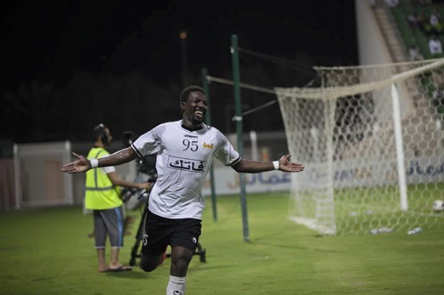 Ein Mann in weißem Hemd und schwarzen Shorts rennt über ein Fußballfeld und breitet die Arme aus, umgeben von Zuschauern, mit einem Torpfosten und einer sitzenden Menge im Hintergrund.