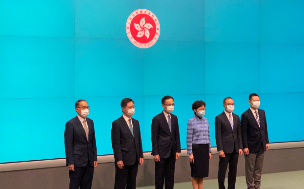 Eine Gruppe von Menschen mit Masken steht nebeneinander vor einer blauen Wand mit einem Logo und nimmt an einer Pressekonferenz in Hong Kong teil.