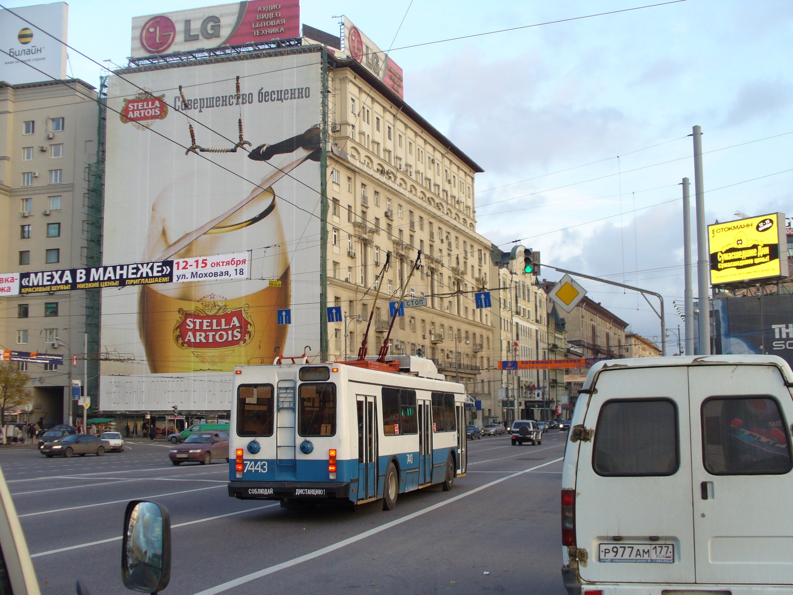 Eine belebte Stadtstraße mit mehreren Fahrzeugen, darunter ein Bus und ein Lieferwagen, gesäumt von Gebäuden, Schildern, Strommasten mit Drähten und Ampeln unter einem bewölkten Himmel.