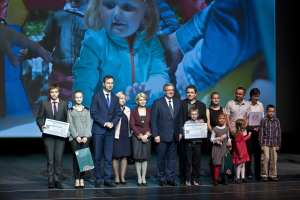 Eine Gruppe von Menschen auf einer Bühne mit Taschen und Urkunden, im Hintergrund ein Bildschirm mit einem Bild eines Kindes und anderen