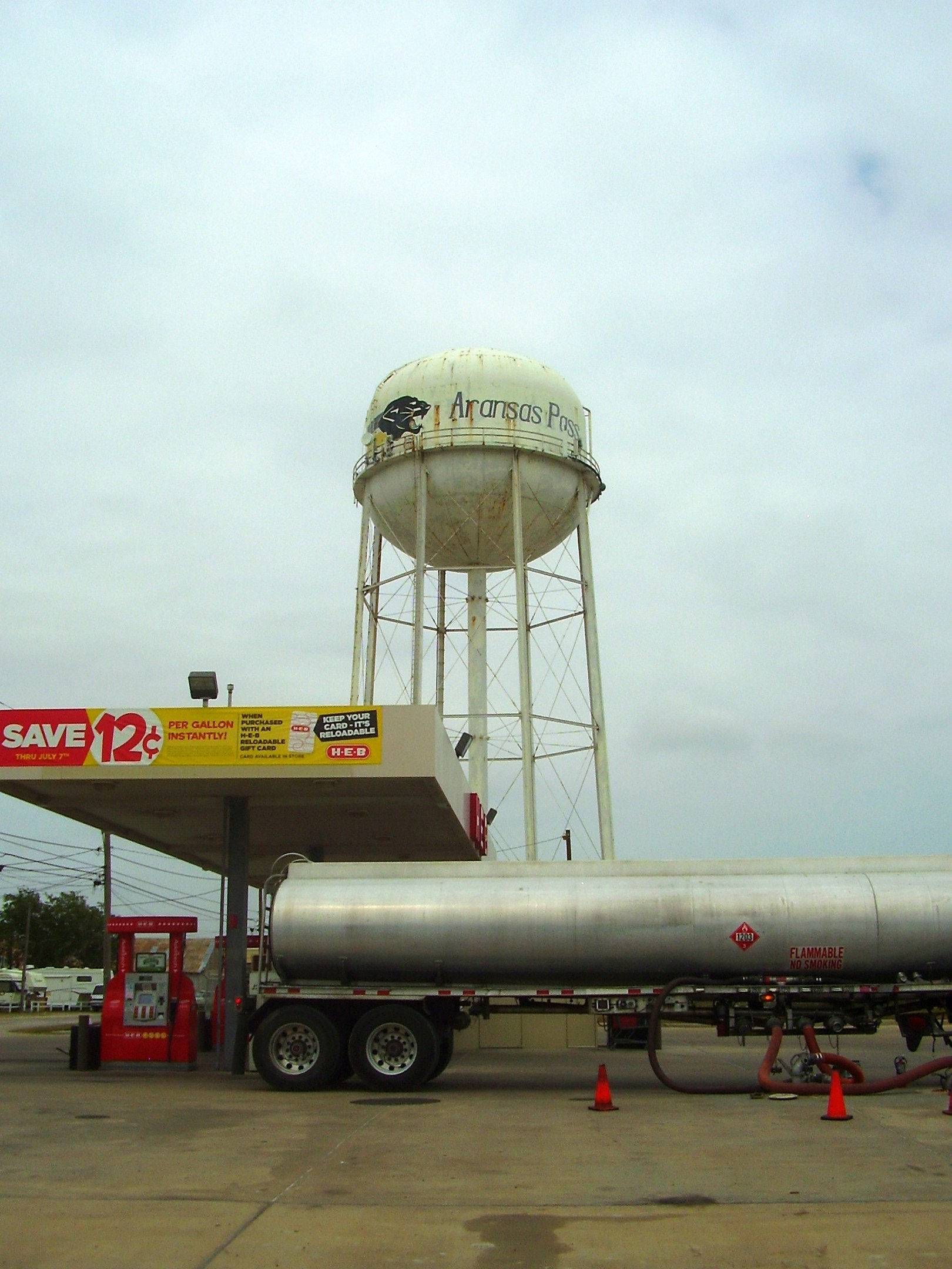 Großer Tanklastwagen vor einer Tankstelle mit Zapfsäule, Absperrgittern und anderen Fahrzeugen auf der Straße, mit einem Wasserturm, Bäumen, Masten, Drähten und einem klaren blauen Himmel im Hintergrund.