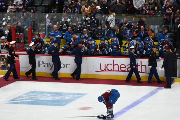 Ein Hockey-Spieler auf dem Eis umgeben von Menschen mit Caps, die Hockey-Stöcke halten, mit Zuschauern im Hintergrund und Tafeln mit der Aufschrift 'Colorado Avalanche vs San Jose Sharks - 3/24/15 NHL Pick, Odds, and Prediction'.
