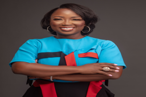 Eine Frau in einem blauen Oberteil und schwarzem Rock, identifiziert als Nana Akufo-Addo, steht mit verschränkten Armen und strahlt Selbstbewusstsein aus.