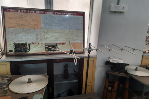 Ein unordentlicher Raum mit verschiedenen Elektronikgeräten, darunter eine beschriftete Platine auf einem Tisch, Metallstangen, eine wandmontierte Schalttafel, ein Fenster und Gegenstände in Schränken, die auf eine computergeschaltete Modus-Stromversorgung hindeuten.