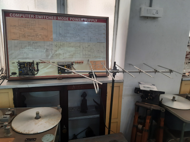 Ein unordentlicher Raum mit verschiedenen Elektronikgeräten, darunter eine beschriftete Platine auf einem Tisch, Metallstangen, eine wandmontierte Schalttafel, ein Fenster und Gegenstände in Schränken, die auf eine computergeschaltete Modus-Stromversorgung hindeuten.