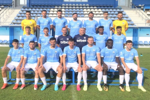 Männer in Sportkleidung stehend und sitzend auf einem Fußballfeld mit Tribünen im Hintergrund.