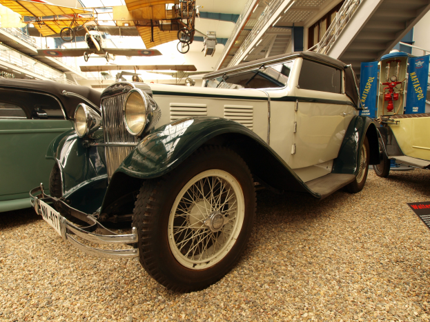 Ein Oldtimer in einem Museum zwischen anderen Fahrzeugen, mit einer Tafel auf der rechten Seite und Flugzeugen an der Decke im Hintergrund.