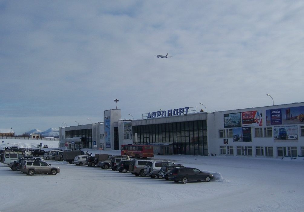 Flughafen-Terminal mit schneebedeckten geparkten Autos im Vordergrund, angrenzende Gebäude mit Fenstern und Beschriftungen, Laternenpfähle, bewölkter Himmel und ein Flugzeug im Flug.
