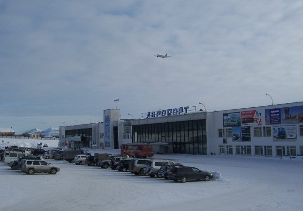 Flughafen-Terminal mit schneebedeckten geparkten Autos im Vordergrund, angrenzende Gebäude mit Fenstern und Beschriftungen, Laternenpfähle, bewölkter Himmel und ein Flugzeug im Flug.