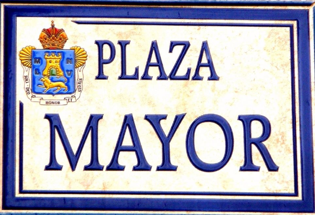 Rechteckige weiße Plakette mit schwarzem Text "Plaza Mayor" in fetter Schrift, blauem Rand und einem zentralen gekrönten Wappen-Logo an einer Gebäudefassade.