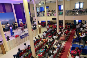 Eine große Gruppe von Menschen, die auf Stühlen in einer Kirche sitzen, mit einigen Individuen auf der Bühne, einem Podium mit einem Mikrofon auf der linken Seite und einer Decke, die mit Lichtern und Lüftern geschmückt ist.